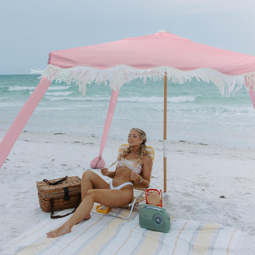 A woman sitting under AMMSUN 6'×6' Gentle Pink Bobo Beach Cabana with Fringe on the beach