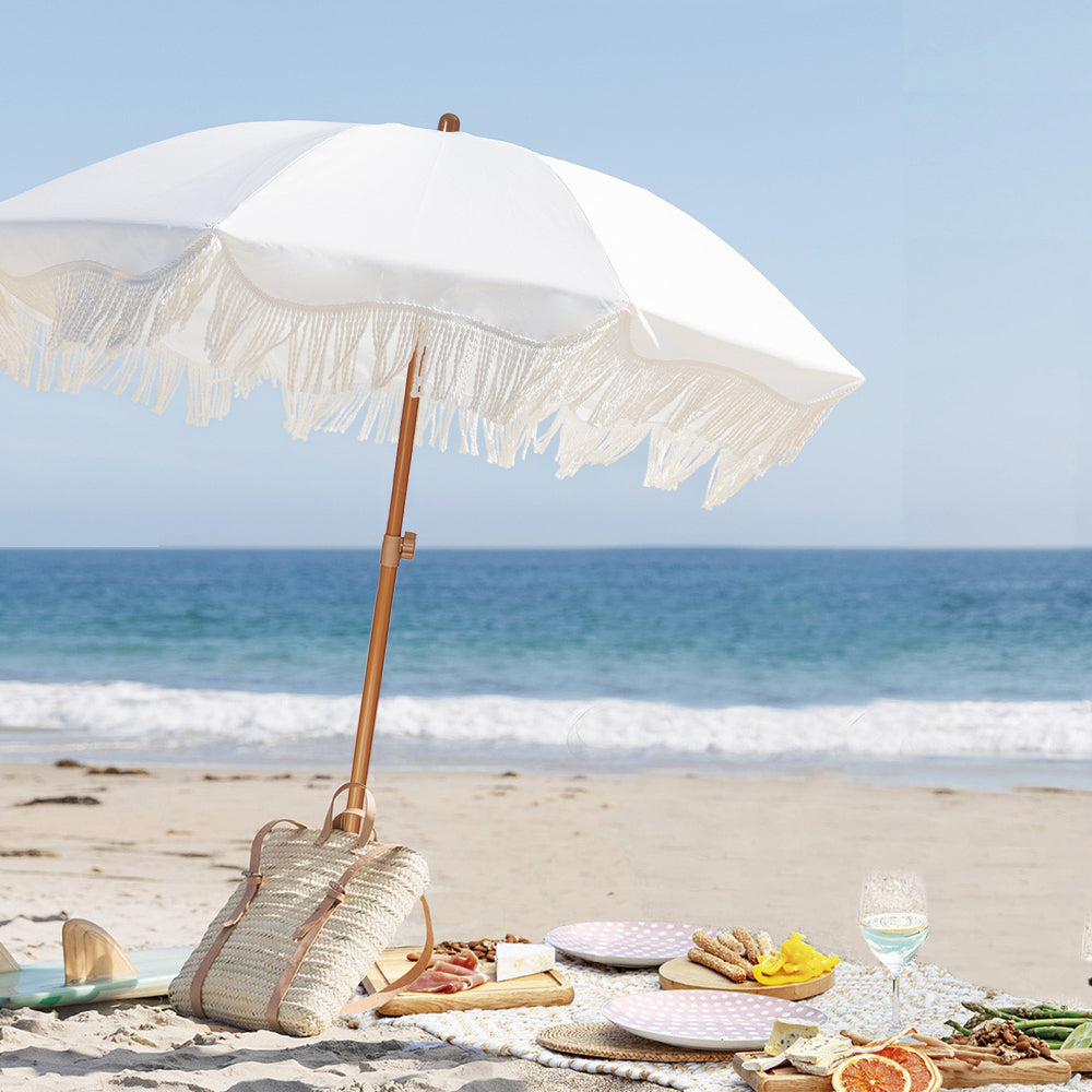 A serene beach scene featuring a picnic blanket shaded by an AMMSUN 6.5ft white beach umbrellas with fringe