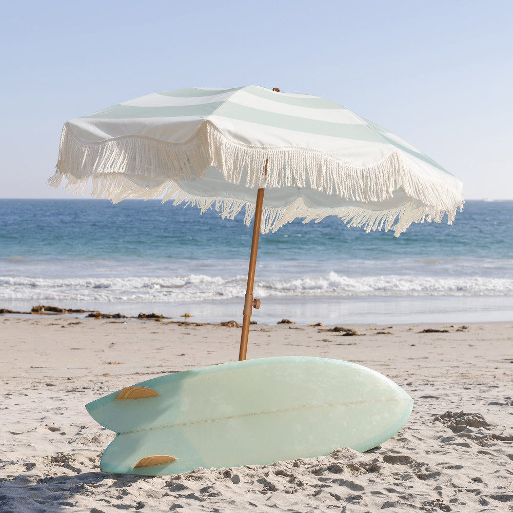 A colorful surfboard positioned on the shore under AMMSUN 6.5ft Vintage Green boho fringe portable beach umbrella