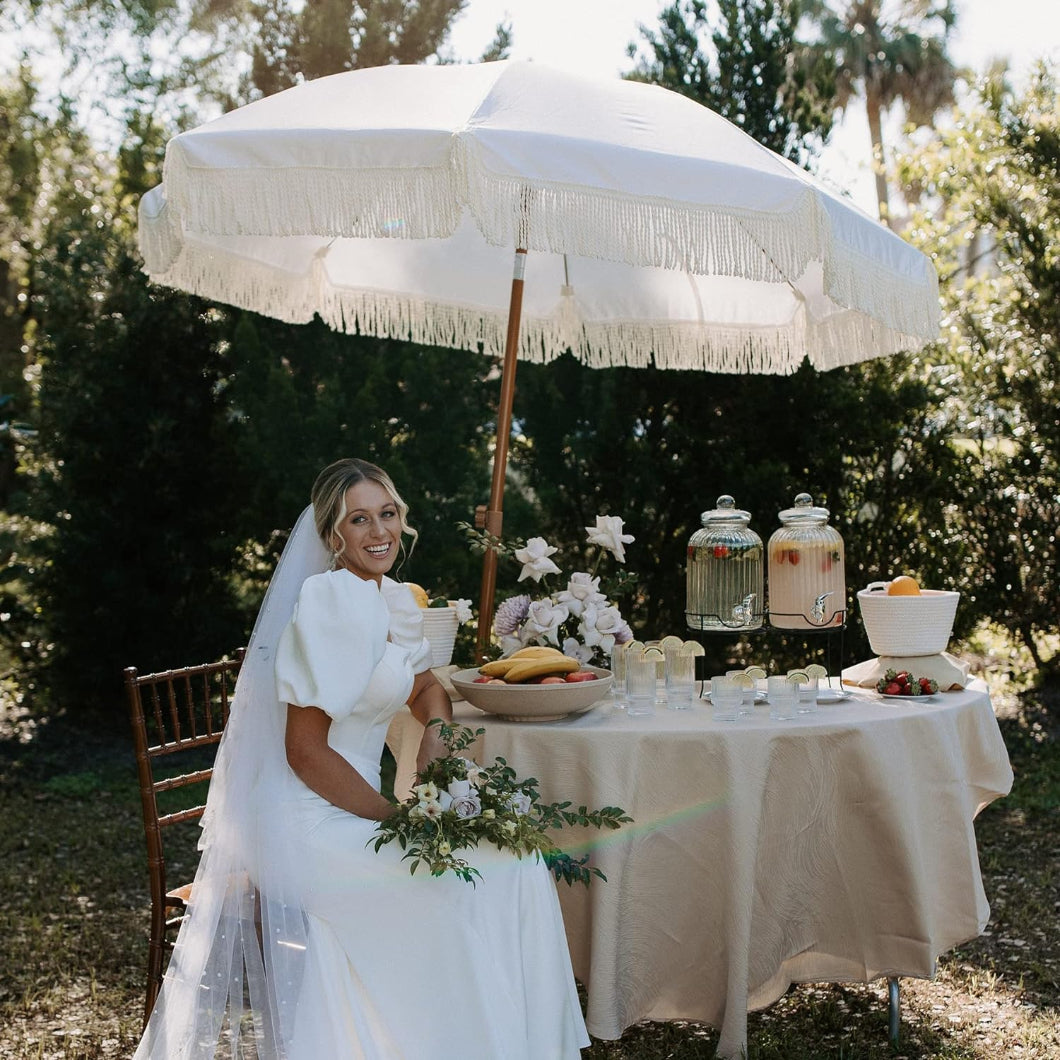 A bride seated at a table with food, sheltered under AMMSUN 7ft elegant cream fringe table umbrellas