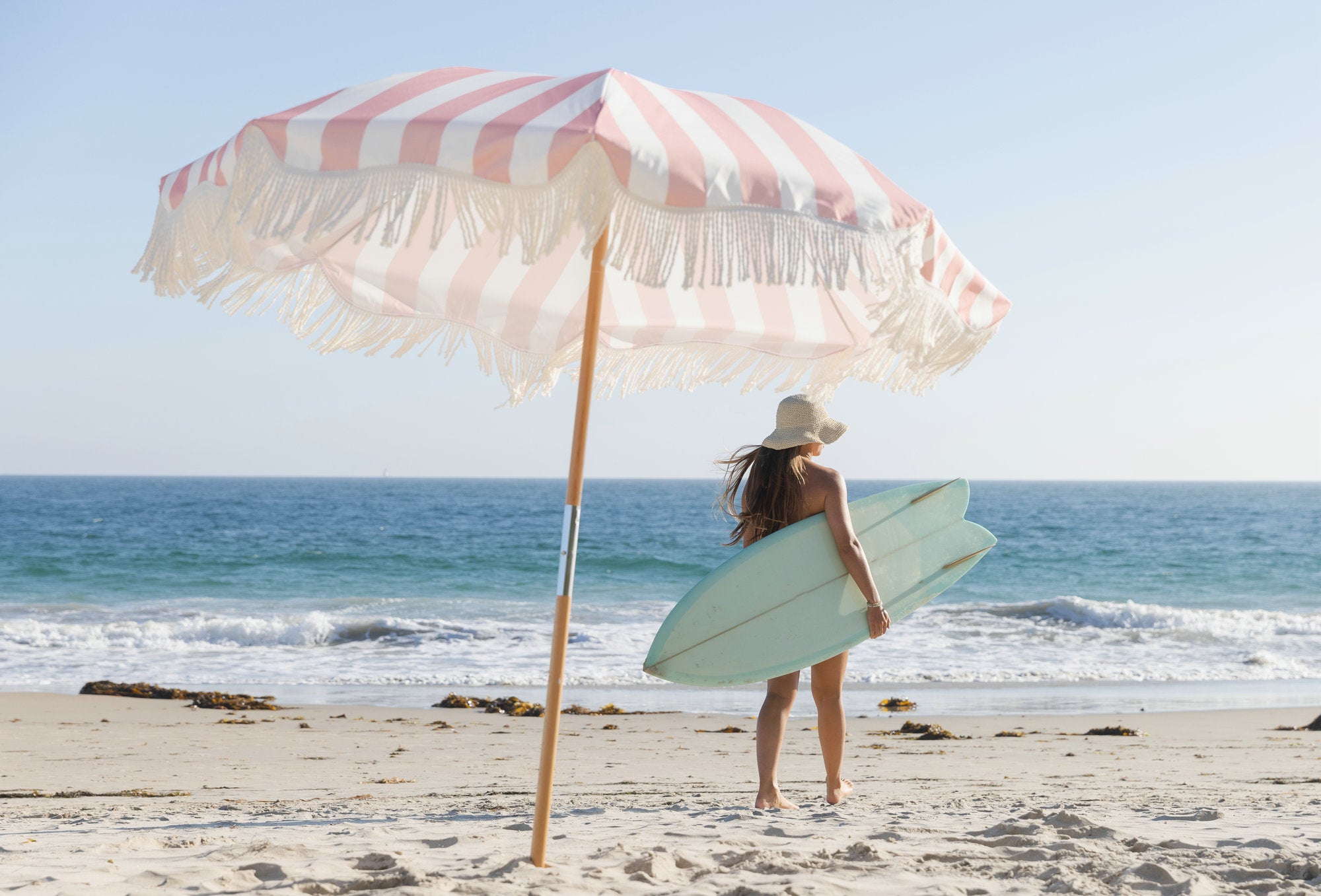 A woman with a surfboard under an AMMSUN 6.5ft pink white outdoor umbrella with fringe on beach 